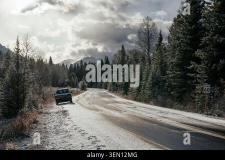 Schneebedeckte Straße schlängelt sich durch einen Wald unter bewölktem Himmel mit einem geparkten Auto auf der linken Seite. Stockfoto