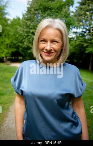 Eine lächelnde Frau mit kurzen weißen Haaren, die ein blaues Oberteil trägt, steht draußen in einem Park. Stockfoto