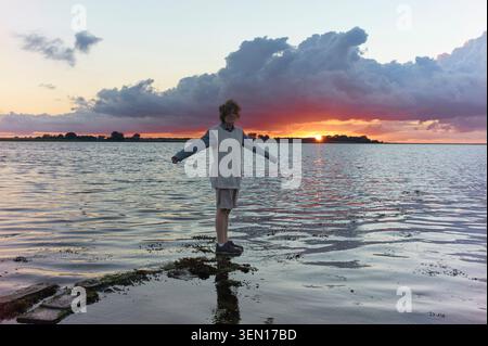 Die Person steht auf einem Felsen in einem See bei Sonnenuntergang mit dramatischen Wolken am Himmel. Stockfoto