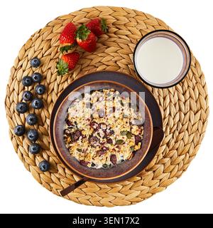 Blick von oben auf Müsli mit Milch in einer Frühstückschüssel, frische Erdbeeren, Heidelbeeren und ein Glas Milch auf gewebtem Tischset. Stockfoto