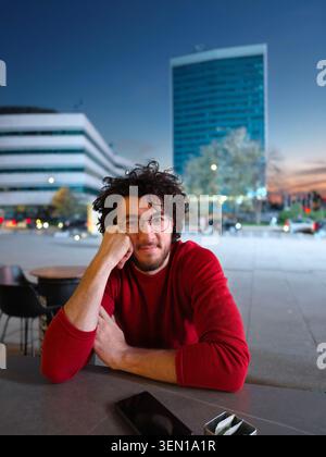 Junger Mann in rotem Pullover, der am Outdoor Cafe Tisch im Urban Evening Portrait sitzt Stockfoto