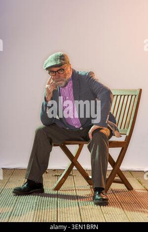 Malachi O'Doherty sitzt auf einem Holzstuhl mit Brille und Mütze. Er posiert nachdenklich, mit einer Hand am Kinn, in einem Studio-Setting mit weichem l Stockfoto