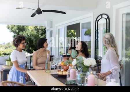 Verschiedene Freundinnen umarmen und plaudern an einem langen Tisch auf der Veranda mit Cupcakes und Champagner Stockfoto
