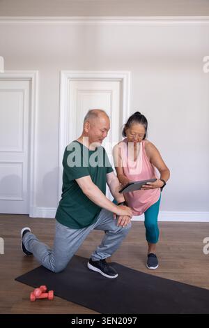Asiatisches Paar kniet mit Tablette, trainiert in Sportbekleidung auf Yogamatte zu Hause mit rosa Kurzhanteln Stockfoto