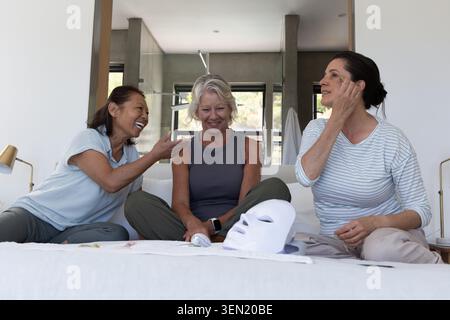 Verschiedene Freundinnen mit Senioren und mittleren Alters, die mit LED-Maske und -Rollen auf dem Bett sitzen Stockfoto