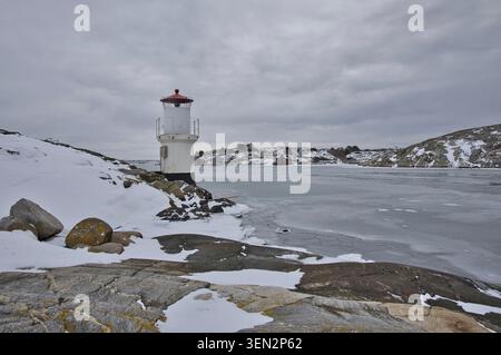 Kleiner weißer Leuchtturm mit rotem Dach an der gefrorenen Küste. Nordisches Meereszeichen in einer winterlichen Landschaft aus Eis und Schnee aus Mollösund, Schweden Stockfoto