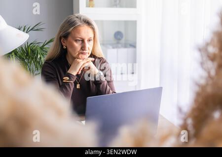 Konzentrierte Geschäftsfrau mittleren Alters, die allein am Schreibtisch mit Laptop arbeitet, denkt und sich auf Aufgaben im modernen Home Office konzentriert. Konzept der Freiberuflichkeit Stockfoto