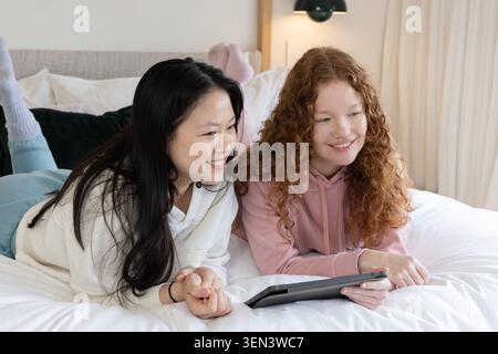 Verschiedene Freundinnen liegen nebeneinander auf dem Bett im Schlafzimmer, schauen sich ein Tablet an und tragen Kapuzenpullover Stockfoto