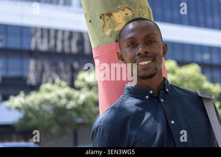 Erwachsener afrikanischer Mann, der sich am rosa umwickelten Baum auf den Bürgersteig lehnt, in dunklem Hemd und Taschenriemen, lächelnd Stockfoto