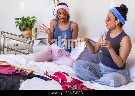 Verschiedene Freundinnen plaudern und trinken Champagner auf dem Bett zu Hause mit Paillettenkleid in der Nähe Stockfoto