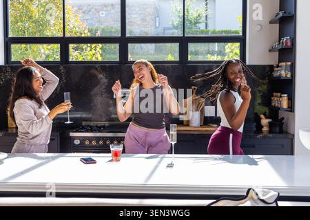 Afroamerikanische Freundinnen in Loungewear tanzen und lachen in der Küche mit Brillentelefon Stockfoto