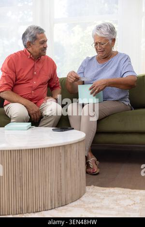Älteres Paar sitzt auf grünem Sofa im Wohnzimmer, Frau liest Karte aus hellblauer Geschenkbox Stockfoto