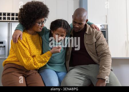 Vielfältige Familie lehnt sich auf die Küchenzeile, Tochter untersucht kleines Spielzeug, Frau trägt Senfstrick Stockfoto