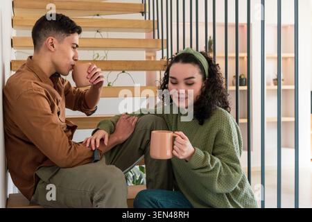 Verschiedene Paare sitzen auf Holztreppen zu Hause, halten passende Tassen und tragen braunes Hemd Stockfoto