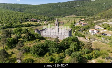 Drohnenansicht der befestigten Dreifaltigkeitskirche in Hrastovlje, Slowenien, umgeben von Feldern, Häusern, Bäumen, und sanfte istrische Hügel. Stockfoto