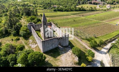 Drohnenblick auf die steinerne befestigte Kirche der Heiligen Dreifaltigkeit in Hrastovlje, Slowenien, mit Blick auf Weinberge, ländliche Straßen und grüne Landschaft. Stockfoto