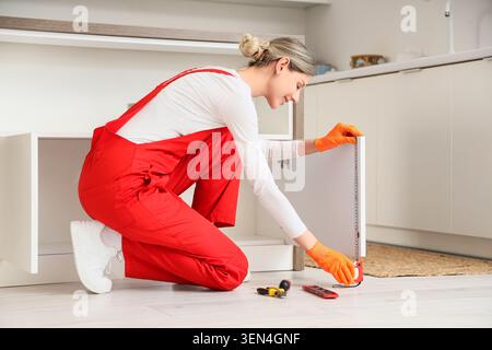 Lächelnde Frau in rotem Overall, die Küchenschrank für Hausrenovierung und Heimwerkerprojekt misst Stockfoto