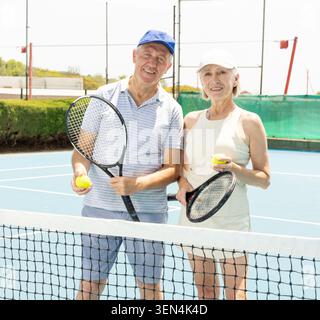 Positiver alter Mann und Frau in Sportkleidung posieren mit Schläger in der Hand neben dem Netz auf einem offenen Tennisplatz Stockfoto