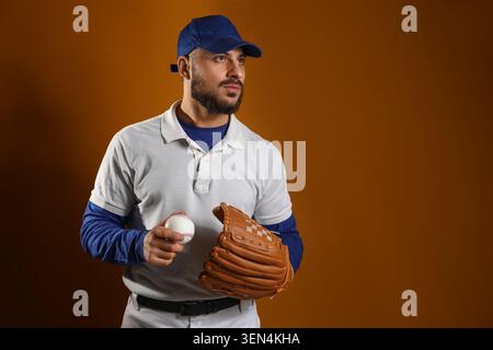 Seriöser Baseballspieler in blauer Kappe und weißem Poloshirt mit Ball und Handschuh, bereit für das Spiel, der Athlet ist konzentriert und bereit für ein Spiel oder ein Training Stockfoto