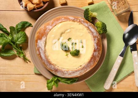 Cremige Brokkoli-Käsesuppe in Sauerteigbrotschale mit frischem Basilikum und Croutons auf Holztisch, einer Seite knuspriger Croutons und frischen Basilikumblättern Co Stockfoto