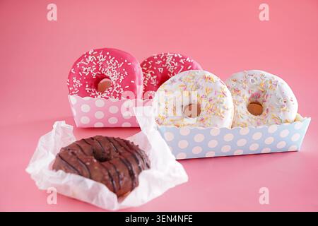 Verschiedene süße Donuts mit bunten Streuseln und Schokoladenglasur in Papierschalen auf rosa Hintergrund, zwei rosa-mattierte Donuts mit weißen Streuseln sind Stockfoto