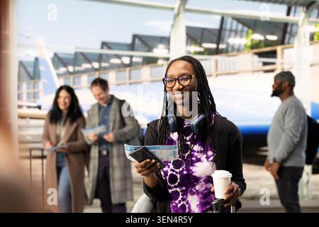 Lächelnde afroamerikanerin am Flughafen mit Kaffeetasse zum Mitnehmen, Smartphone und Bordkarte, die begeistert ist, den Urlaub zu beginnen. Fröhlicher Touristenspaziergang im belebten Flughafen mit Gepäck und Reiseutensilien Stockfoto