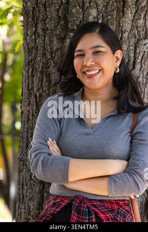 Erwachsene Frau, die sich im Park gegen einen Baum lehnt und lächelt, Arme gekreuzt, graugestricktes Karo trägt Stockfoto