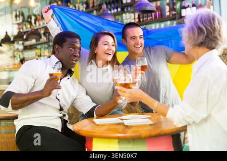 Fröhliche multirassische Männer und Frauen feiern den Teamsieg, winkende Flagge der Ukraine in Bar Stockfoto
