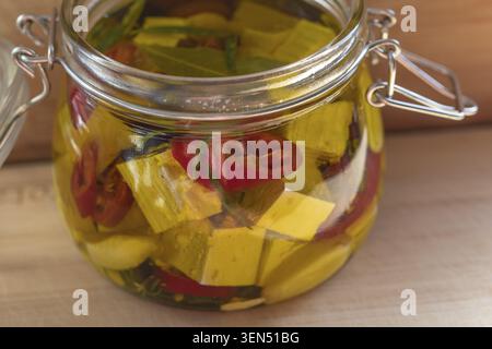 Aus nächster Nähe ein Glasgefäß mit offenem Drahtbügel, gefüllt mit marinierten gelben Feta- oder Tofu-Würfeln, Lorbeerblättern, Chili und Rosmarin in Olivenöl Stockfoto