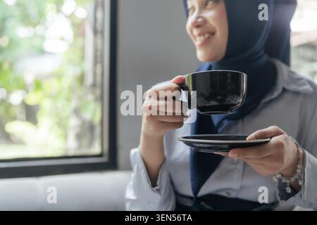 Lächelnde Frau im Hijab, die eine Tasse und Untertasse in der Nähe eines Fensters in einem hellen Café hält Stockfoto