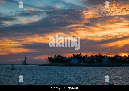 Das kleine Passagiersegelboot überquert das Meereswasser in der Nähe der dunklen Silhouette der Key West Island unter dem bewölkten Himmel während des Sommersonnenverganges. Stockfoto