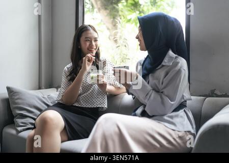Zwei Frauen unterhalten sich und genießen Drinks auf einem Sofa am hellen Fenster in einem entspannten Wohnzimmer Stockfoto