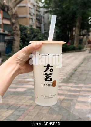 TAIPEH, TAIWAN - 4. MAI 2026: Eine Hand hält eine aus Taiwan markierte Teetasse mit Daming-Markenzeichen auf einem von Bäumen gesäumten Bürgersteig in Taiwan, mit Bokeh aus Wohngebäuden und Grün im Hintergrund. Stockfoto