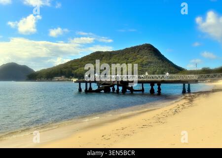 Ein Sandstrand trifft auf ruhiges blaues Wasser, während sich ein Metallpier über das Meer erstreckt, wo Menschen entlang der sonnendurchfluteten Struktur spazieren. Stockfoto