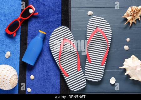 Die Grundausstattung für den Sommerurlaub am Strand liegt flach mit gestreiften Flipflops, roter Sonnenbrille, Sonnencreme und Muscheln auf blauem Handtuch Stockfoto