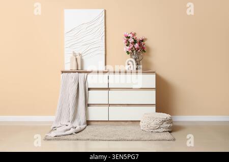 Moderne, minimalistische Kommode mit weißen Schubladen und Holzplatte, dekoriert mit Kunstblumen und gemütlicher Decke in einem neutralen Zimmer Stockfoto