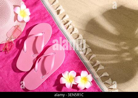 Sommer-Strandurlaub mit rosafarbenen Flipflops, Strohhut, Sonnenbrille und Frangipani-Blumen auf Sand und Handtuch Stockfoto