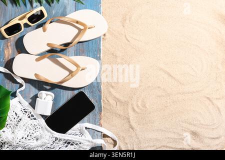Grundausstattung für Sommerurlaub am Strand mit Flipflops, Sonnenbrille, Smartphone und Ohrstöpseln auf Sand und Holz Stockfoto
