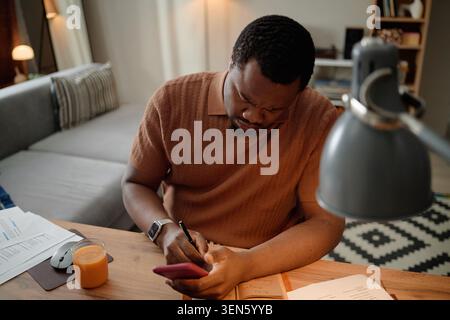 Schwarzer Mann, der das Smartphone benutzt, während er zu Hause arbeitet Stockfoto
