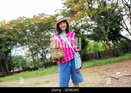 Frau Hut Korb Park Baum rosa Hemd Denim Lächeln im Freien, fröhliche Frau trägt Strohhut, die gewebten Korb im grünen Park mit Baum hält, rosa Hemd, Stockfoto