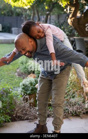 Afroamerikanischer Vater und schulpflichtige Tochter spielen Huckepack im Hinterhof in der Nähe von Terrassenstühlen Stockfoto