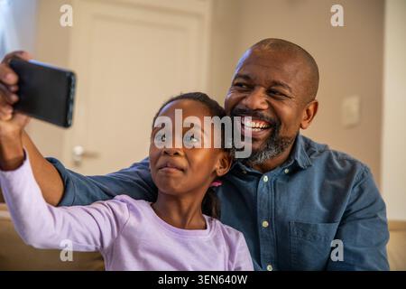 Afroamerikaner Vater und Kind Mädchen machen Selfie auf der Couch zu Hause und halten ein schwarzes Smartphone Stockfoto