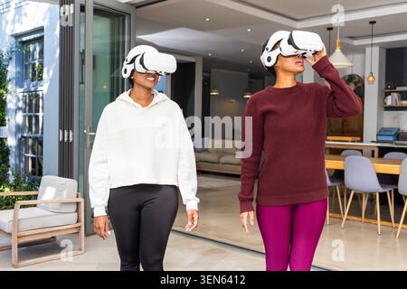 Verschiedene Freundinnen, die zusammen mit weißen VR-Headsets im offenen Wohnbereich zur Terrasse laufen Stockfoto