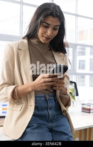 Indianerin lehnt sich an den weißen Schreibtisch, trägt einen beigefarbenen Blazer und benutzt Smartphone mit Laptop Stockfoto