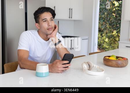 Afroamerikanischer Mann sitzt auf der Marmor-Kücheninsel und hält ein Smartphone, eine Obstschale aus Holz in der Nähe Stockfoto