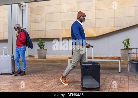 Wandermann in blauem Pullover, orangefarbenen Schuhen, die schwarze Hülle entlang des überdachten Gehwegs ziehen, mit Kopfhörern Stockfoto