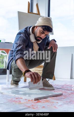 Mann mit Mütze, der sich auf bemaltem Tropfentuch im Studio hockt, mit gekerbter Kelle zur Texturplatte Stockfoto