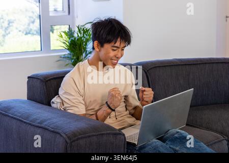 Asiatischer Mann feiert, während er Laptop auf dunkelgrauem Sofa zu Hause in der Nähe des Fensters Armband sichtbar Stockfoto