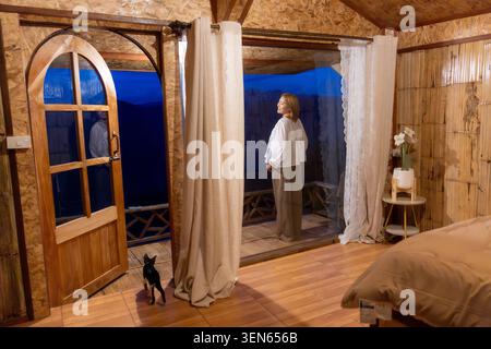 Eine ruhige Frau steht nachts auf dem Balkon eines Holzkabinenzimmers und blickt in die dunkle Dämmerung, während eine kleine Hauskatze in der Nähe einer offenen Bogentür mit weißem C sitzt Stockfoto