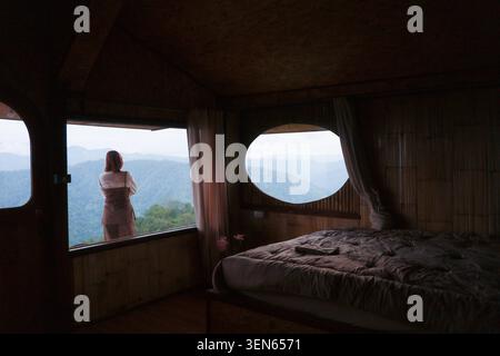 Eine nachdenkliche Frau steht vor einer rustikalen Holzhütte mit Blick durch das Fenster auf den nebeligen Bergblick und genießt den friedlichen Morgen vom gemütlichen dunklen Schlafzimmer in Stockfoto
